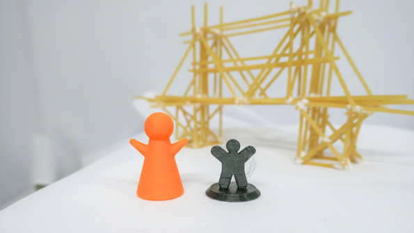 Två 3D-printade plastfigurer står framför en konstruktion av spagetti och lim.