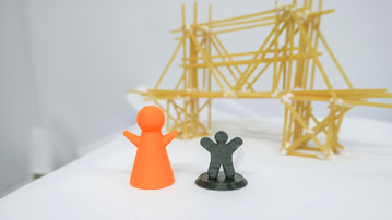 Två 3D-printade plastgubbar står framför en konstruktion av spagetti och lim.