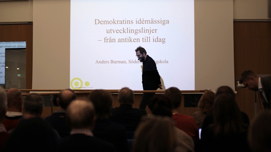 En man föreläser inför en publik och står framför en projektorduk som visar texten "Demokratins idémässiga utvecklingslinjer, från antiken till idag"